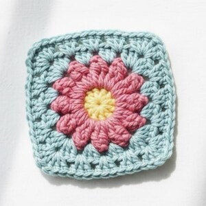 May include: A square, crocheted granny square featuring a central flower design. The flower has a yellow center, surrounded by pink petals. The square's border is a light blue color, creating a vibrant, handmade aesthetic.