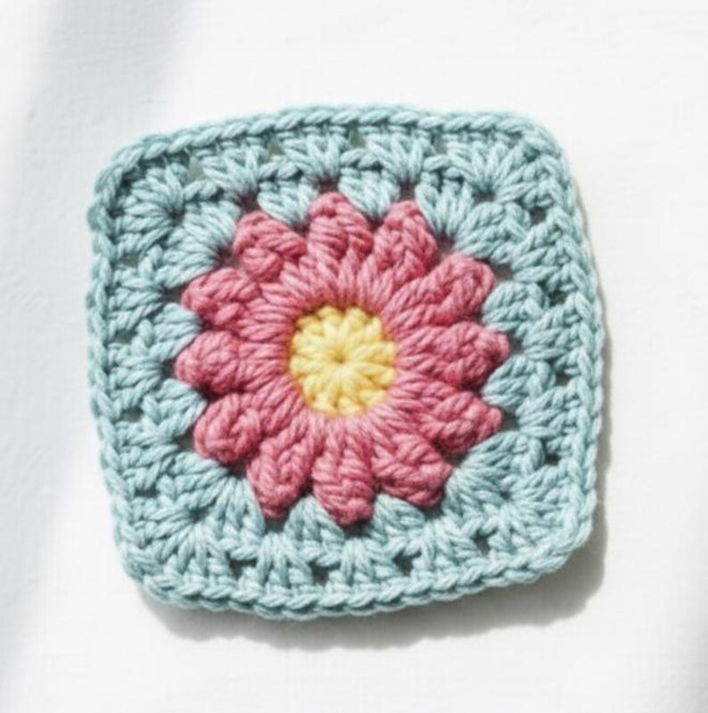 May include: A square, crocheted granny square featuring a central flower design. The flower has a yellow center, surrounded by pink petals. The square's border is a light blue color, creating a vibrant, handmade aesthetic.