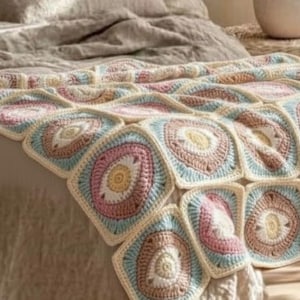 May include: A handmade crochet blanket featuring a repeating square pattern. Each square has a central design in shades of pink, yellow, and brown, surrounded by a light blue and cream border. The blanket is draped over a bed.