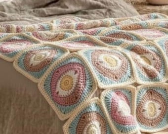 May include: A handmade crochet blanket featuring a repeating square pattern. Each square has a central design in shades of pink, yellow, and brown, surrounded by a light blue and cream border. The blanket is draped over a bed.