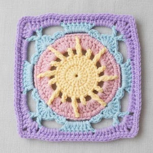 May include: A square, crocheted sunburst design in pastel colors. The center features a yellow sun shape surrounded by a pink circle. Light blue and lavender crochet work frames the design, creating a textured, handmade appearance. This item is suitable for various craft projects.