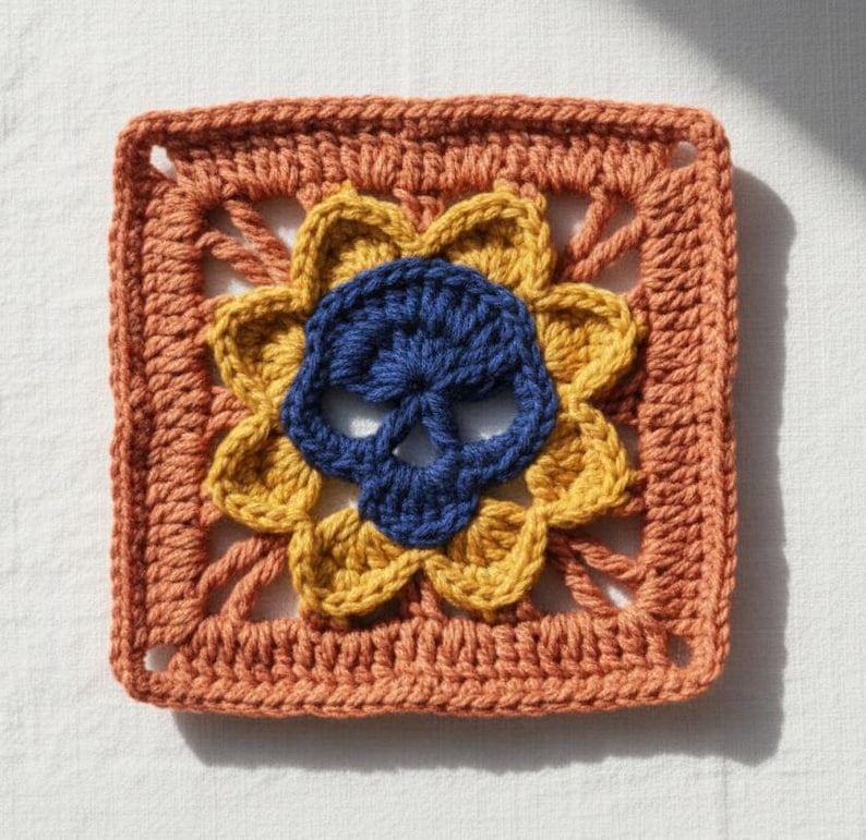 May include: Crocheted square featuring a blue skull at the center, surrounded by yellow petals and a rust-colored border. The handmade piece has a textured appearance, suitable for various craft projects or decorative use.