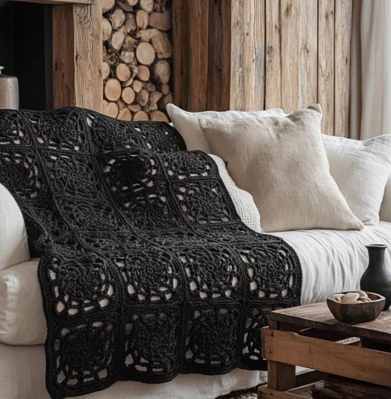 May include: A dark gray crocheted blanket draped over a white sofa. The blanket features a square pattern with floral designs. Two beige pillows rest against the back of the sofa. A wooden side table holds a vase and a wooden bowl.