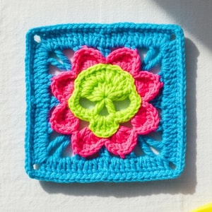May include: A square, crocheted item featuring a neon green skull at the center, surrounded by a pink flower design. The skull and flower are set against a bright blue square background. The item is handmade.