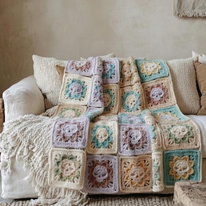 Puede incluir: Una manta de ganchillo en colores pastel con un diseño de calavera. La manta, en tonos crema, lavanda y verde azulado, está sobre un sofá blanco con cojines de tonos neutros. El diseño de calavera se repite en los bloques cuadrados.