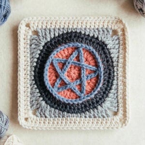 Puede incluir: Un elemento cuadrado tejido a ganchillo con un diseño central de pentagrama. El pentagrama es azul, sobre un círculo de color coral, rodeado de anillos concéntricos grises y negros. El borde exterior es de color crema, con pequeños recortes cuadrados en cada esquina.