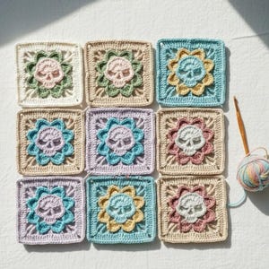May include: Nine crocheted squares in various pastel colors, each featuring a skull design at the center, surrounded by a floral pattern. A crochet hook and a ball of yarn are visible, suggesting a handmade craft project.