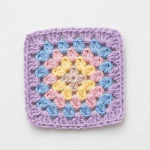 May include: A square, crocheted granny square in pastel colors. The center is a light brown, surrounded by yellow, pink, and blue petals. The outer border is a lavender color, with a textured stitch pattern.