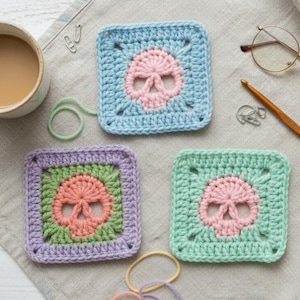 May include: Three crocheted square coasters with skull designs in pink, blue, green, and purple. A cup of coffee, a crochet hook, and glasses are on a neutral linen surface. The coasters are handmade.