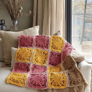 May include: A handmade crochet blanket in shades of pink, yellow, and tan. The blanket is made of square motifs with a spiral design in the center. The blanket is draped over a beige sofa, with a neutral background.
