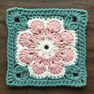May include: A square, handmade crochet piece featuring a central flower design. The flower has white and pink petals, surrounded by a teal border with leaf-shaped accents. The square is approximately 15 cm by 15 cm.