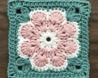 May include: A square, handmade crochet piece featuring a central flower design. The flower has white and pink petals, surrounded by a teal border with leaf-shaped accents. The square is approximately 6 inches by 6 inches.