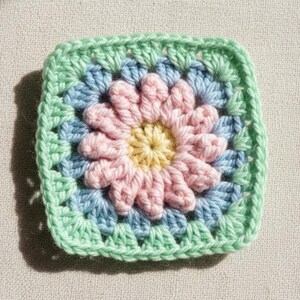 May include: A square, handmade crochet piece featuring a floral design. The center is yellow, surrounded by pink petals, light blue, and a green border. The yarn appears soft and the stitches are tight, creating a textured, colorful square.