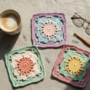 May include: A collection of colorful, crocheted square coasters with sunburst designs. The coasters are in shades of pink, green, and purple, with a cup of coffee and eyeglasses in the background. The coasters are handmade.