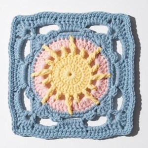 May include: A square, crocheted sunburst design in a granny square pattern. The center features a yellow sun with pink rays, surrounded by a light blue border. The handmade piece is suitable for various craft projects.