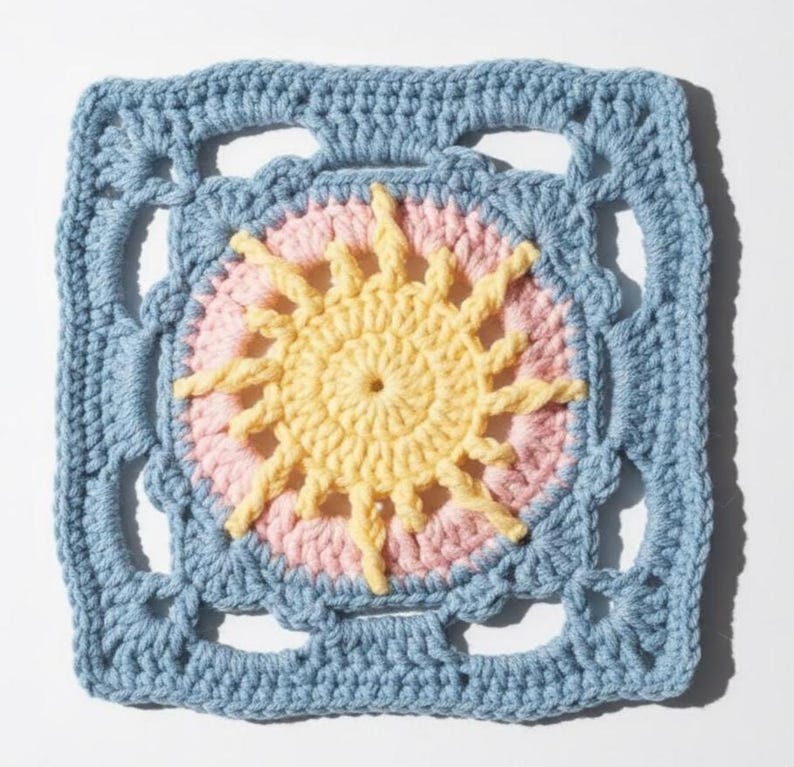 May include: A square, crocheted sunburst design in a granny square pattern. The center features a yellow sun with pink rays, surrounded by a light blue border. The handmade piece is suitable for various craft projects.