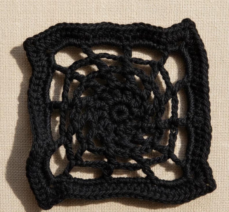 May include: A square, black crochet doily with an open, web-like design. The doily features a spiral center and a solid, textured border. The intricate pattern is visible against a neutral background, highlighting the handmade quality of the piece.