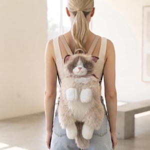 Op de afbeelding: Een pluche rugzak in de vorm van een pluizige ragdoll-kat met lange beige en bruine vacht, witte pootjes en een witte kanten kraag. De kat heeft blauwe ogen, een roze neus en een lange, harige staart. Voorzien van verstelbare beige schouderbanden.