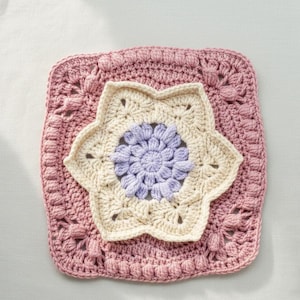 May include: A handmade crochet square in shades of pink, cream, and lavender. The square features a central flower design with a lavender center and cream petals, surrounded by a pink border with textured details. This item is perfect for home decor.