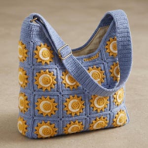 Pode incluir: Uma bolsa de ombro azul de croch&ecirc; com um padr&atilde;o repetido de girass&oacute;is amarelos e brancos. A bolsa tem uma al&ccedil;a longa e uma fivela dourada. O forro interno &eacute; de cor bege claro.
