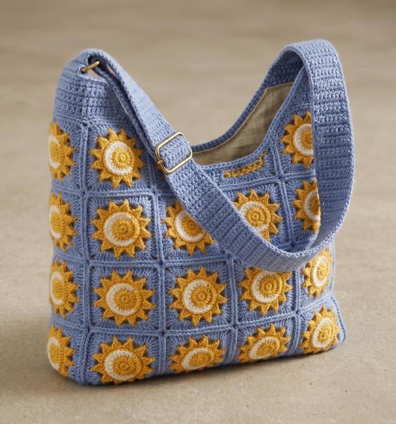 Pode incluir: Uma bolsa de ombro azul de croch&ecirc; com um padr&atilde;o repetido de girass&oacute;is amarelos e brancos. A bolsa tem uma al&ccedil;a longa e uma fivela dourada. O forro interno &eacute; de cor bege claro.