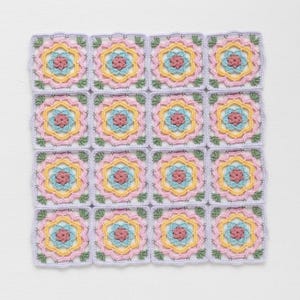May include: A handmade crochet blanket featuring sixteen square motifs. Each square has a floral design with pink, yellow, and blue accents, surrounded by a lavender border. The blanket is a soft, textured piece.