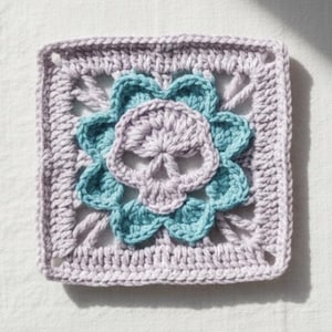 May include: A crocheted square featuring a skull design. The skull is lavender, surrounded by teal flower petals, and set against a lavender square background. The handmade item is likely a decorative element or part of a larger craft project.