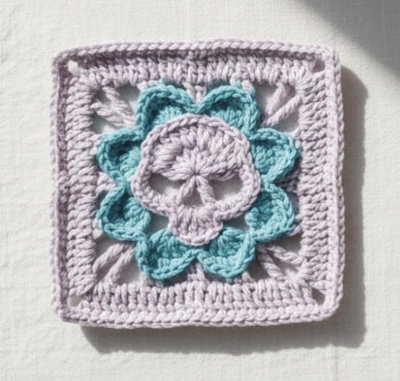 May include: A crocheted square featuring a skull design. The skull is lavender, surrounded by teal flower petals, and set against a lavender square background. The handmade item is likely a decorative element or part of a larger craft project.