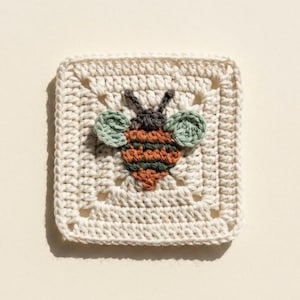 May include: A square, off-white crochet piece featuring a detailed bee applique. The bee has a brown and orange body with green wings and a dark gray head. The crochet work has a textured appearance, with a repeating pattern around the edges.