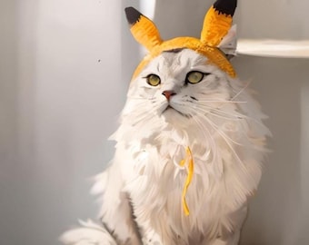 May include: A fluffy white cat wearing a yellow and black fox ear hat. The cat has green eyes and a yellow ribbon around its neck. The cat is sitting on a wooden surface.