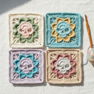 May include: Four crocheted squares with skull and flower designs in pastel shades. Each square features a skull motif at the centre, encircled by a floral pattern. Colours include cream, blue, pink, and tan. A crochet hook and yarn are visible.