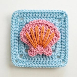 May include: A square, crocheted piece featuring a light blue background with a pink seashell design. The seashell has orange detailing. The square is a granny square, a common crochet technique. The piece is handmade.