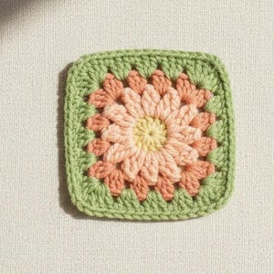 May include: A square, crocheted granny square in a floral design. The center is yellow, surrounded by peach and coral petals, and framed by a green border. The square is set against a neutral background.