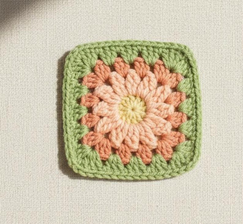 May include: A square, crocheted granny square in a floral design. The center is yellow, surrounded by peach and coral petals, and framed by a green border. The square is set against a neutral background.
