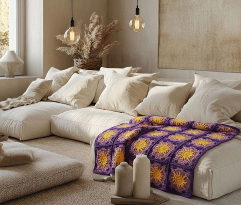 May include: A cozy living room scene featuring a modular sofa with cream-colored cushions and pillows. A vibrant, hand-crocheted blanket in purple and yellow adds a pop of color. Decorative elements include pendant lights and dried plants.