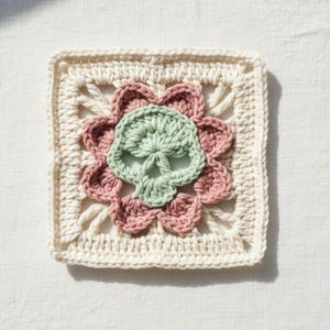 May include: A square, crocheted motif featuring a green skull at the center, surrounded by a pink flower design. The skull and flower are set against a cream-colored background, with a square border. The piece is handmade.