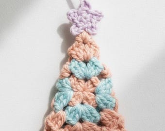 May include: A handmade crochet Christmas tree ornament. The tree is made of peach, light blue, and brown yarn, with a lavender star on top. The ornament has a loop for hanging. A festive, handcrafted decoration.