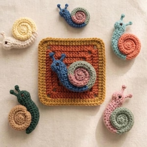 May include: A selection of crocheted snail ornaments in various colours. The snails have spiral shells and are in shades of blue, pink, green, and yellow. One snail is attached to a square, crocheted coaster with a brown and yellow border.