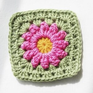 May include: A square, handmade crochet piece featuring a central pink flower with a yellow center, surrounded by a green square border. The crochet stitches are visible, creating a textured effect. This item is likely a decorative element or part of a larger craft project.