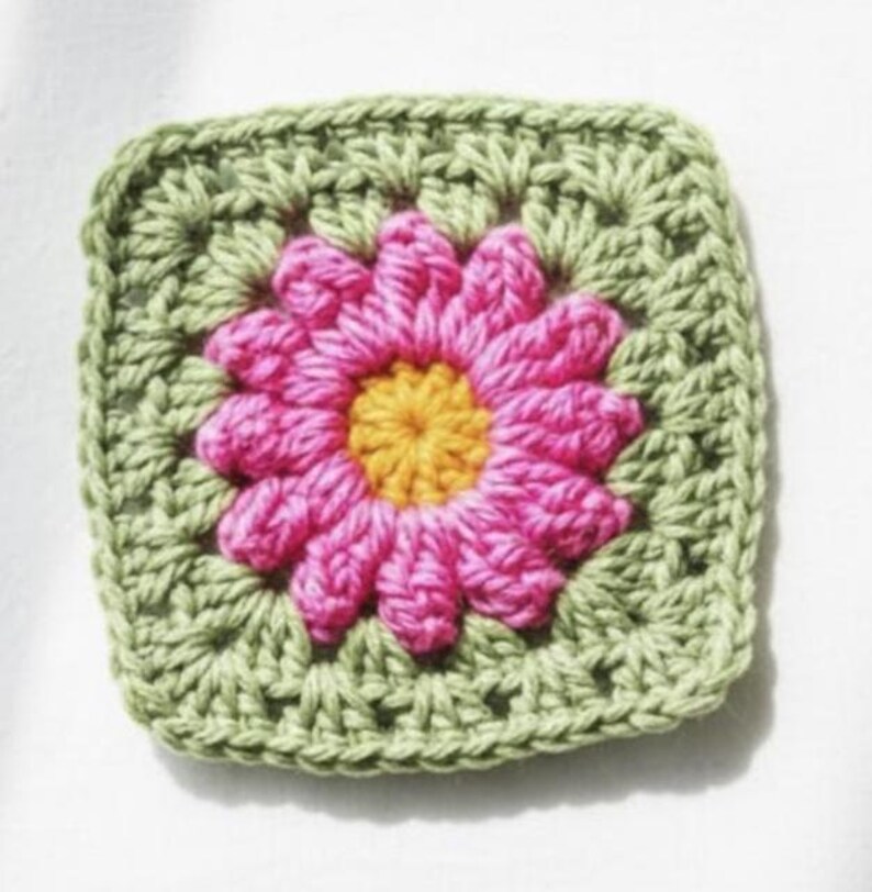 May include: A square, handmade crochet piece featuring a central pink flower with a yellow center, surrounded by a green square border. The crochet stitches are visible, creating a textured effect. This item is likely a decorative element or part of a larger craft project.