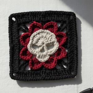 May include: A square, crocheted motif featuring a skull design. The skull is light gray, surrounded by red flower petals, and framed by a black square. The design is centered on a white surface.