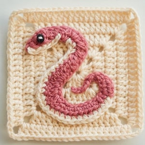 Op de afbeelding: Een gehaakte vierkant met een roze en wit slangenmotief. De slang is het centrale element, met een gedetailleerd, getextureerd uiterlijk. De vierkant is lichtbeige van kleur, met een getextureerd patroon en een vierkante vorm.