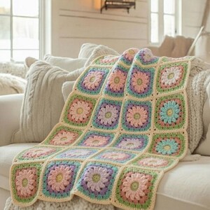 May include: A handmade crochet blanket featuring a floral pattern. The blanket is made of squares in pastel colors, including pink, purple, green, and blue, with a cream-colored border. It is draped over a white sofa, creating a cozy and inviting look.