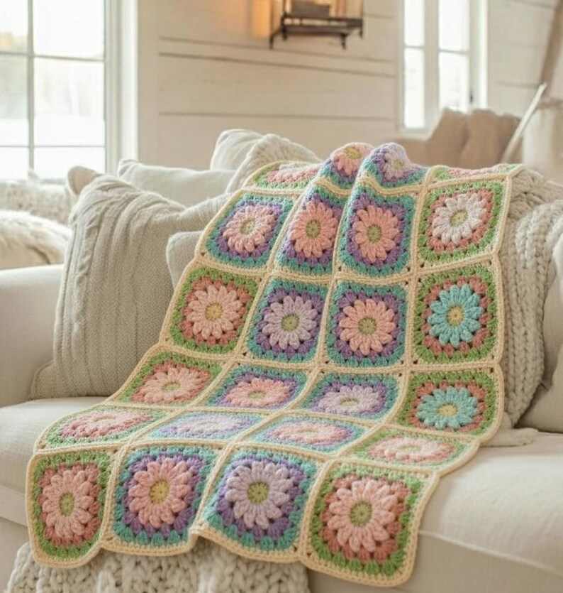 May include: A handmade crochet blanket featuring a floral pattern. The blanket is made of squares in pastel colors, including pink, purple, green, and blue, with a cream-colored border. It is draped over a white sofa, creating a cozy and inviting look.