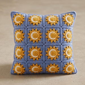 Pode incluir: Uma almofada quadrada de croch&ecirc; com fundo azul e um padr&atilde;o de s&oacute;is amarelos e brancos. A almofada &eacute; feita de quadrados individuais, cada um com um desenho de sol. A almofada tem aproximadamente 40 cm quadrados.