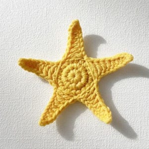 May include: A handmade, yellow crocheted starfish. The five-pointed star has a textured surface and a circular center. The bright yellow color and detailed stitching create a charming, nautical-themed decorative item.