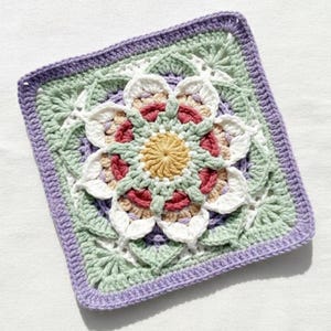 May include: A square, handmade crochet piece featuring a central floral design. The flower has a yellow center, surrounded by white, pink, and beige petals. The square is bordered by light green, lavender, and purple yarn.
