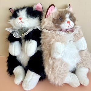 May include: Two plush cat toys with long faux fur. One cat is black and white with a green bow and lace collar. The other cat is tan, brown, and white with a pink bow and lace collar. Both have pink ears, embroidered faces, and white paws.