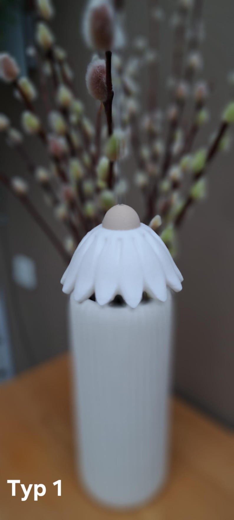 May include: A white ceramic diffuser with a daisy-shaped top and a cylindrical base. The text "Typ 1" is visible at the bottom. The background features a blurred arrangement of pussy willow branches.