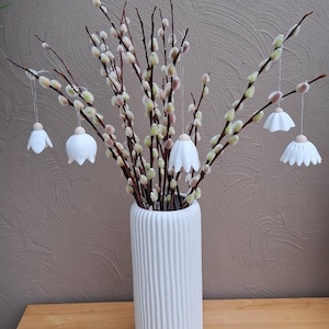 May include: A white, ribbed ceramic vase holds pussy willow branches with hanging white flower ornaments. The arrangement sits on a wooden surface against a textured brown wall. Decorative and seasonal.